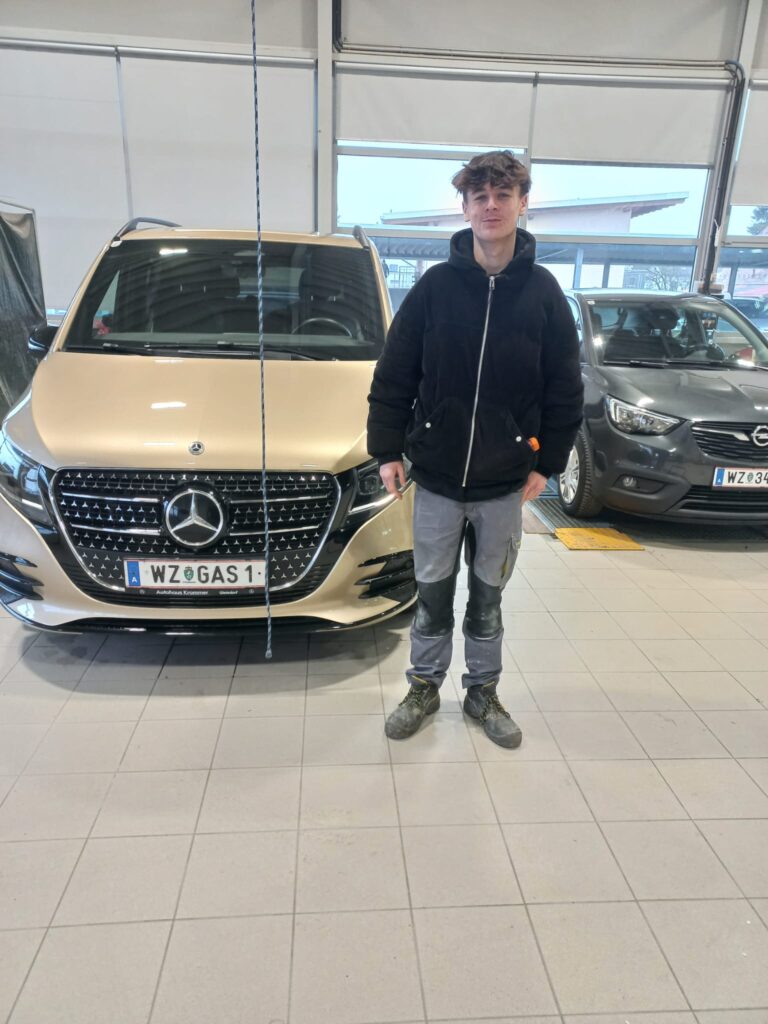 Junger Mann in Werkstatt vor goldener Mercedes-V-Klasse und grauem Opel