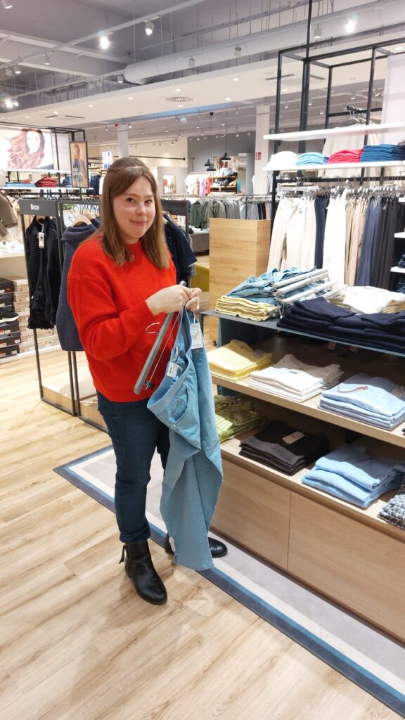 Frau im roten Pullover hält hellblaue Jeans in einem Bekleidungsgeschäft in Wien